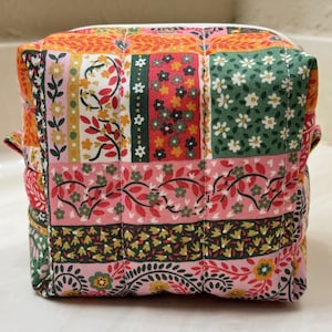 May include: A square, quilted cosmetic bag with a patchwork design. The bag features a variety of floral patterns in shades of pink, green, orange, and yellow. It has a white zipper closure and small fabric loops on the sides.