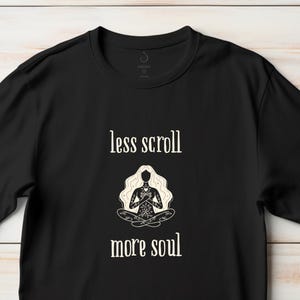 May include: Black t-shirt with the text "less scroll more soul" and a graphic of a person meditating. The graphic is in white and beige. The t-shirt is made of a soft material.