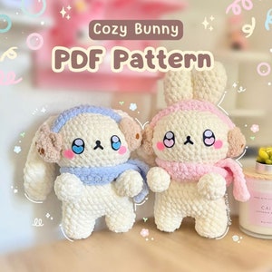 Könnte beinhalten: Zwei gehäkelte Hasen-Plüschtiere mit weißem Fell, blauen und rosa Schals und gestickten Gesichtern. Die Plüschtiere sitzen auf einem Tisch mit einem rosa und weißen Hintergrund. Der Text "Cozy Bunny PDF Pattern" ist über den Plüschtieren zu sehen.
