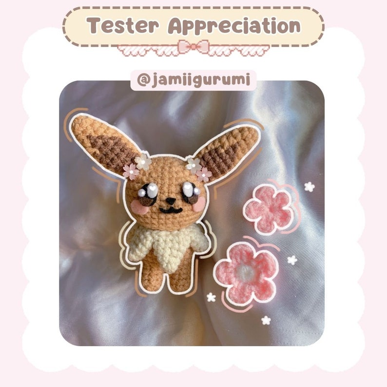 May include: A crocheted Eevee plush toy with brown and white fur, pink cheeks, and pink flower accents. The toy is sitting on a pink satin fabric with pink flower embellishments. The text "Tester Appreciation" and "@jamiigurumi" are displayed above the toy.