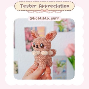 May include: A crocheted Eevee plush toy in a light brown color with a white collar. The toy is being held in a person's hand. The background is blurred and features a pink and white floral pattern.