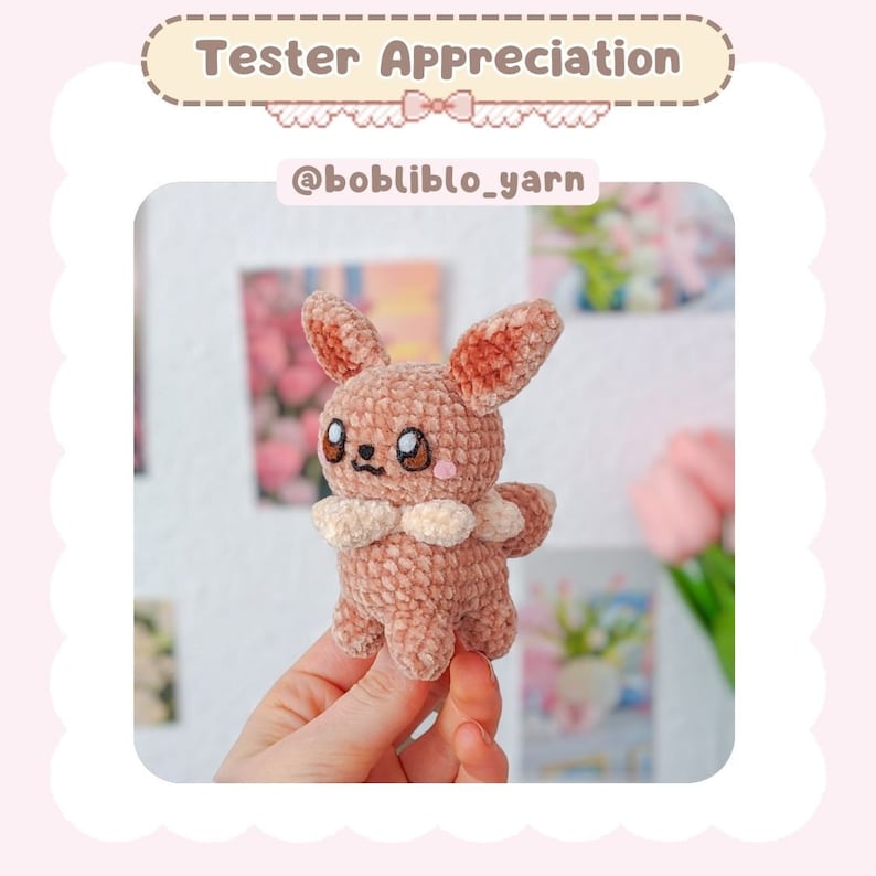 May include: A crocheted Eevee plush toy in a light brown color with a white collar. The toy is being held in a person's hand. The background is blurred and features a pink and white floral pattern.