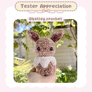 May include: A brown and pink crocheted Eeveelution plush toy with black eyes. The toy is being held in a person's hand.