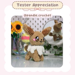 May include: A crocheted Eevee plush toy in brown and white with a white collar. The toy is sitting on a table with a mirror reflecting the toy.
