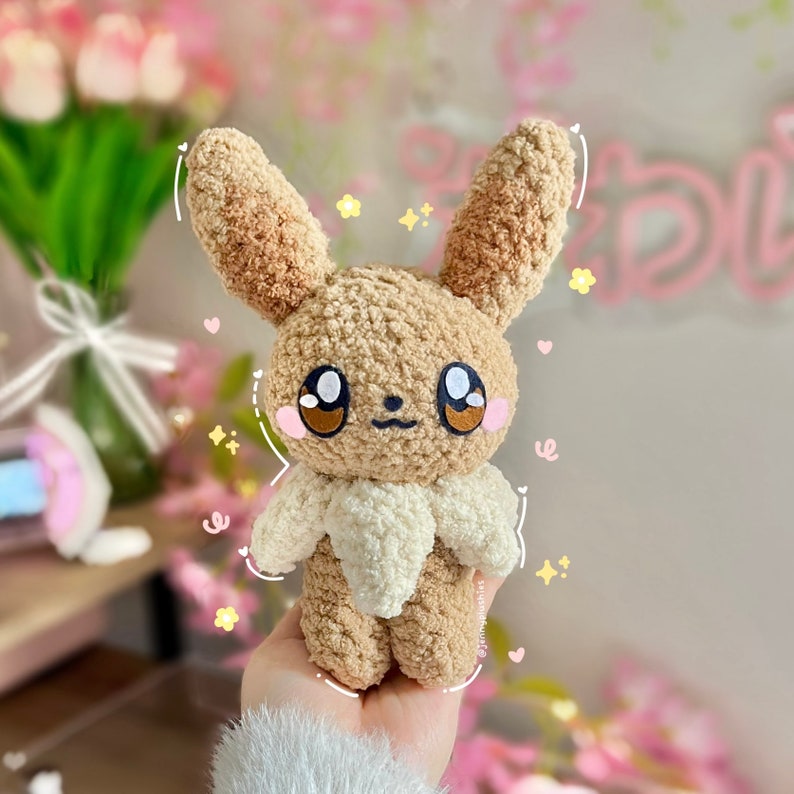 May include: A brown and white crocheted plush toy resembling a Pokemon character. The toy has large brown eyes, a small brown nose, and a white fluffy collar.