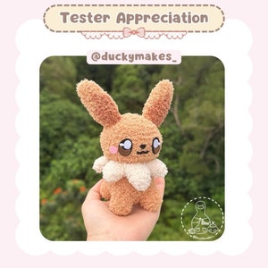 May include: A brown and white knitted Eevee plush toy with big eyes. The toy is being held in a hand against a green background. The text "@duckymakes_" is visible in the image.