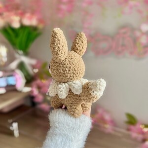 May include: A brown and white crocheted Eevee plush toy with a fluffy tail. The toy is sitting on a person's hand and is facing away from the camera.