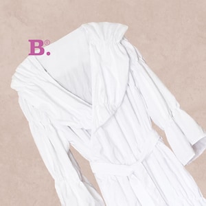 May include: A white dress with a V-neckline, long sleeves, and a belted waist. The dress has a flowing design with ruffled details on the sleeves and neckline. The dress is made of a soft, lightweight fabric.