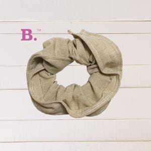 May include: A beige fabric scrunchie with a textured surface. The scrunchie is tied in a knot and is ready to be worn.