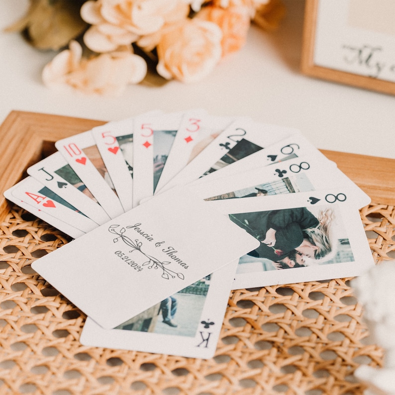 May include: A deck of playing cards with a custom design featuring a photo of a couple and the text "Jessica & Thomas 05.21.2023". The cards are fanned out on a woven wicker surface.