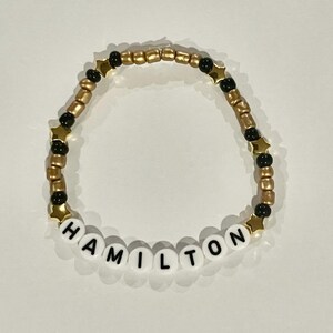 Peut inclure: Un bracelet en perles dorées et noires avec le mot "HAMILTON" écrit en lettres blanches.