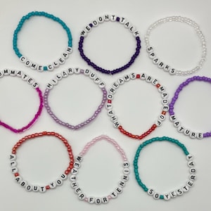 May include: Ten circular beaded bracelets featuring white letter beads with black text. Phrases include COME CLEAN, WE DONT TALK, SPARKS, ROOMMATES, HILARY DUFF, WHAT DREAMS ARE MADE OF, MATURE, ALL ABOUT YOU, WEATHER FOR TENNIS, and SO YESTERDAY.