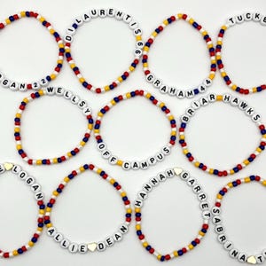 Op de afbeelding: Een verzameling armbanden met kralen met witte letterkralen die namen en zinnen spellen. De armbanden hebben rode, gele en blauwe kralen. Sommige armbanden bevatten cijfers of hartvormige kralen. Op een witte achtergrond.