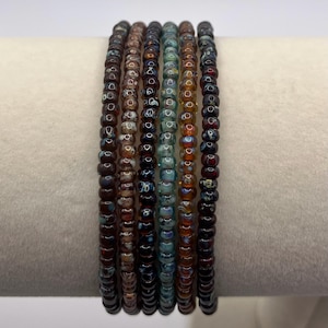 Puede incluir: Una pila de siete pulseras de cuentas. Las pulseras presentan una variedad de colores, incluyendo marrón, verde azulado y naranja. Las cuentas son pequeñas y redondas, y las pulseras están apiladas sobre una superficie blanca.
