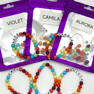 Puede incluir: Tres bolsas moradas resellables con etiquetas blancas que dicen VIOLET, CAMILA y AURORA, cada una con una pulsera de cuentas multicolor. Debajo, tres pulseras adicionales presentan cuentas con letras blancas que forman los nombres EMMA, ANNA y OLIVIA.