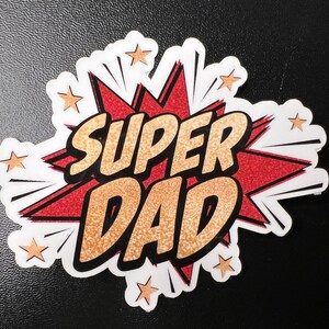 Op de afbeelding: Een rode en witte stripboek-stijl grafiek met gouden glitter accenten. De grafiek zegt "SUPER DAD" in vetgedrukte zwarte letters.