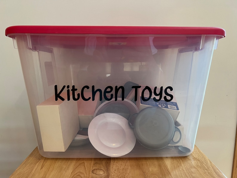 Pode incluir: Uma caixa de armazenamento de pl&aacute;stico transparente com uma tampa vermelha. A caixa est&aacute; cheia de v&aacute;rios brinquedos de cozinha, incluindo um prato branco, uma panela preta e uma caixa etiquetada como "Pieces 99". A caixa est&aacute; etiquetada como "Kitchen Toys" em letras pretas.