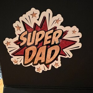 Op de afbeelding: Een witte sticker met een rode en gouden stripboek-stijl grafiek die "SUPER DAD" zegt met gouden sterren rond de grafiek.
