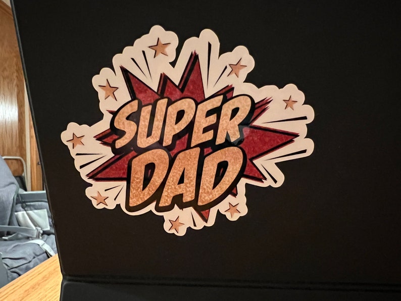 Op de afbeelding: Een witte sticker met een rode en gouden stripboek-stijl grafiek die "SUPER DAD" zegt met gouden sterren rond de grafiek.