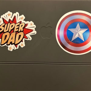 Pu&ograve; includere: Un tablet grigio con due adesivi. Un adesivo dice "SUPER DAD" in un font in stile fumetto con accenti rossi e dorati. L'altro adesivo &egrave; uno scudo di Capitan America rosso, bianco e blu.