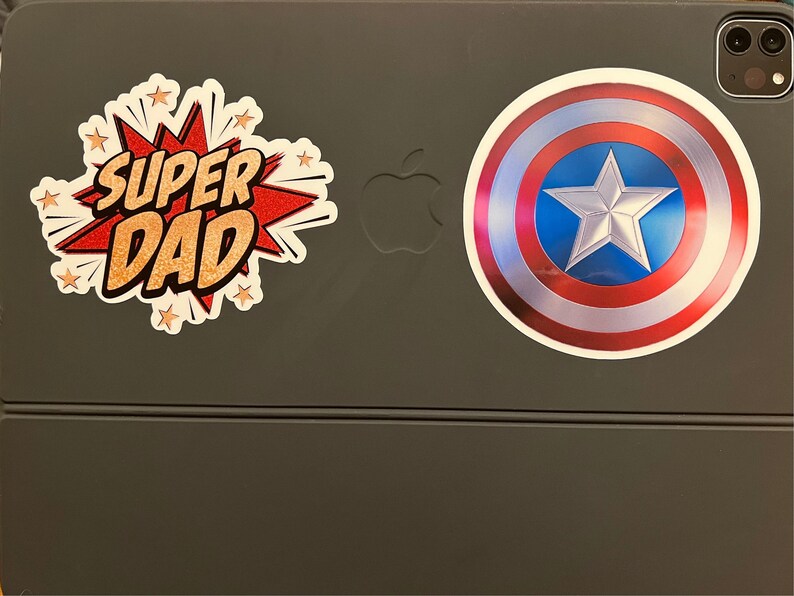 Pu&ograve; includere: Un tablet grigio con due adesivi. Un adesivo dice "SUPER DAD" in un font in stile fumetto con accenti rossi e dorati. L'altro adesivo &egrave; uno scudo di Capitan America rosso, bianco e blu.