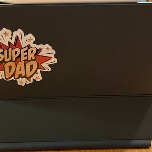 Op de afbeelding: Een zwarte iPad-hoes met een sticker waarop "SUPER DAD" staat in een rood en gouden comic-lettertype.
