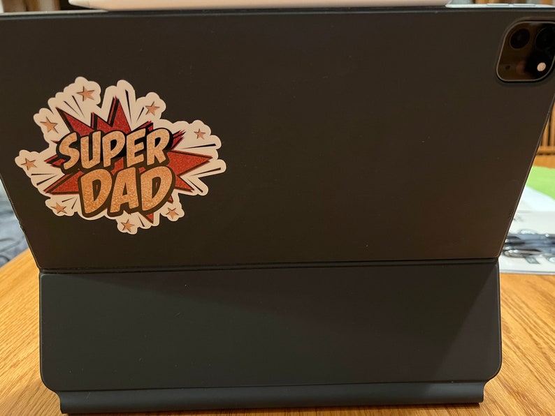 Op de afbeelding: Een zwarte iPad-hoes met een sticker waarop "SUPER DAD" staat in een rood en gouden comic-lettertype.