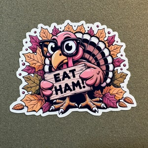 Pu&ograve; includere: Un tacchino dei cartoni animati con gli occhiali e che tiene un cartello con su scritto "Eat Ham!" Il tacchino &egrave; circondato da foglie autunnali.