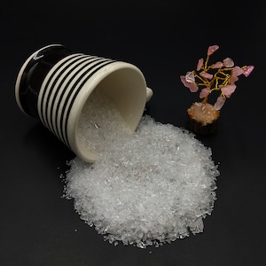 Könnte beinhalten: Eine schwarz-weiß gestreifte Keramiktasse verschüttet klare kristallartige Perlen auf eine schwarze Oberfläche. Rechts von der Tasse steht ein kleiner rosa Kristalldrahtbaum.