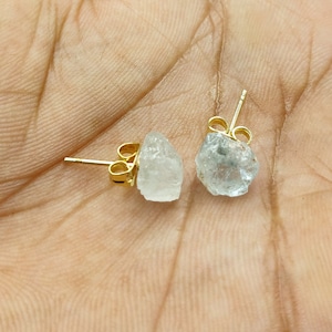 May include: A pair of gold-colored stud earrings, each featuring a rough-cut, light blue-gray gemstone. The earrings are set on gold posts with butterfly clasps. The gemstones have a natural, irregular shape.