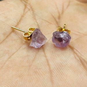 Pode incluir: Um par de brincos de pressão de ametista. Cada brinco apresenta uma pedra de ametista bruta, roxa clara, montada em um pino dourado. Os brincos são exibidos em uma superfície marrom clara.