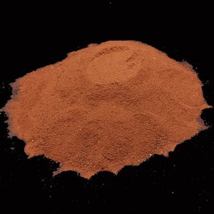 May include: A mound of reddish-brown powder, finely ground, set against a black backdrop. The powder's texture appears slightly irregular, with subtle colour and density variations. The shape is a rounded heap, indicating a granular material.