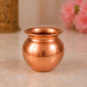 May include: A small, polished copper pot with a rounded body and a wide, flared rim. The metallic surface features a warm, reddish-brown hue with a smooth, reflective finish. The vessel has a short, narrow neck that transitions into a bulbous base.