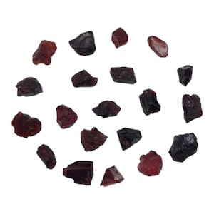 May include: A collection of rough, dark red garnet gemstones. The stones vary in size and shape, with a mix of deep burgundy and black hues. The gemstones are scattered against a white background, showcasing their natural, unpolished texture.