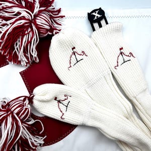 May include: Golf club head covers in cream-colored knit with a red flag design embroidered on each. The set includes a black club identifier with a white "X" and red and white pom-poms.