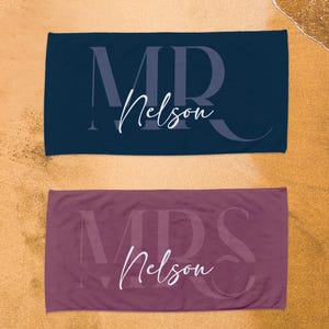 May include: Two rectangular beach towels, one dark navy blue and one mauve, each featuring a large, faint, capitalized monogram in the background and the white cursive text "Nelson" printed prominently across the center of the fabric.