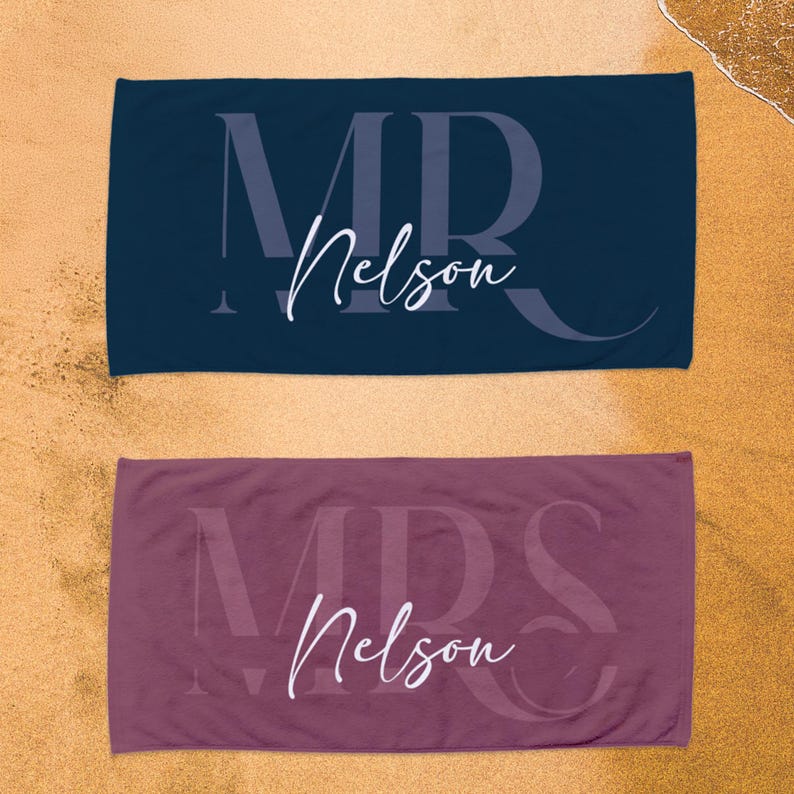 May include: Two rectangular beach towels, one dark navy blue and one mauve, each featuring a large, faint, capitalized monogram in the background and the white cursive text "Nelson" printed prominently across the center of the fabric.