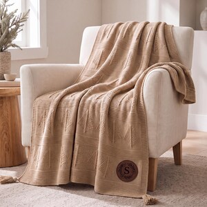 May include: A tan knitted throw blanket featuring a repeating pattern of triangular tree motifs and horizontal textured bands. The blanket includes a circular brown leather patch with the letter S embossed in the center and tassel ends.