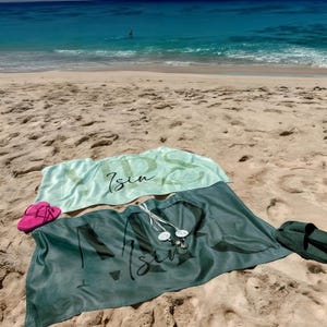 May include: Two rectangular beach towels in light mint green and dark forest green, each featuring the text "KIDS" and "Isin" in a stylized script font. A pair of bright pink flip-flops and dark green slides are placed on the sand beside them.