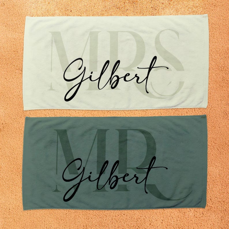 May include: Two rectangular towels, one light cream and one dark sage green. Each towel features a large, faint, capitalized 'MRS' or 'MR' in the background, overlaid with the name 'Gilbert' written in a prominent black cursive script font.