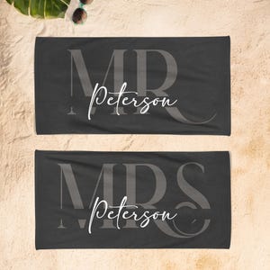 May include: Two rectangular charcoal gray beach towels featuring large, dark gray block-letter monograms of MR and MRS. Each towel is personalized with the white cursive script text Peterson centered over the monograms in a clean, modern font style.