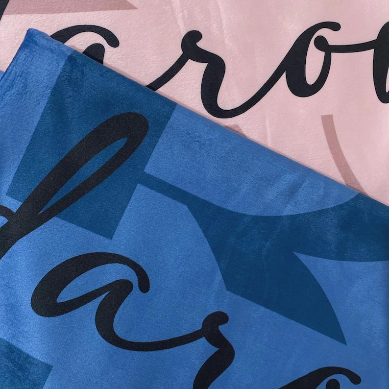 May include: A soft, plush fabric blanket featuring a layered design. The top layer is a light pink material with black cursive lettering. The bottom layer is a deep blue fabric with a darker blue geometric pattern and black cursive lettering.
