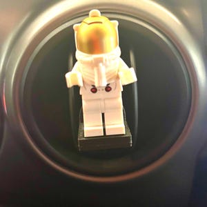 Könnte beinhalten: Eine weiße Lego-Astronauten-Minifigur mit goldenem Helm ist an einem schwarzen Lüftungsgitter im Auto befestigt.