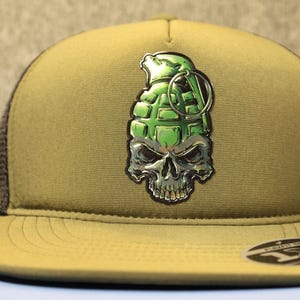 May include: A khaki-colored trucker hat with a green and silver grenade skull emblem. The hat has a curved brim and a mesh back. The emblem features a green grenade with a skull design.