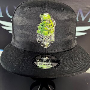 以下が含まれることがあります： 黒と緑の迷彩柄のベースボールキャップで、9FIFTY SNAPBACKのラベルが付いています。キャップには、銀色の骸骨と手榴弾のデザインが施されています。