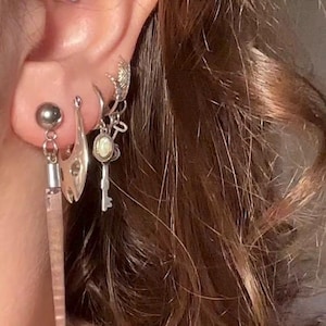Pode incluir: Uma coleção de brincos prateados em múltiplos furos na orelha, incluindo um pingente retangular longo, fino e rosa translúcido, uma forma metálica curva, uma pequena argola com um pingente de chave e um pequeno brinco de tarraxa.