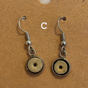 Puede incluir: Un par de pendientes colgantes plateados. Cada pendiente presenta un centro circular beige con un patr&oacute;n radial, rodeado por un marco de metal plateado. Los pendientes se muestran en una tarjeta marr&oacute;n.