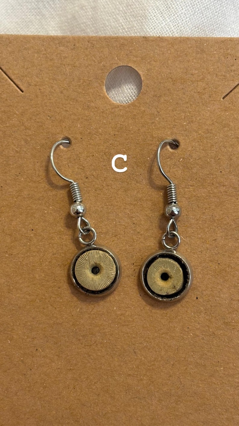 Puede incluir: Un par de pendientes colgantes plateados. Cada pendiente presenta un centro circular beige con un patr&oacute;n radial, rodeado por un marco de metal plateado. Los pendientes se muestran en una tarjeta marr&oacute;n.