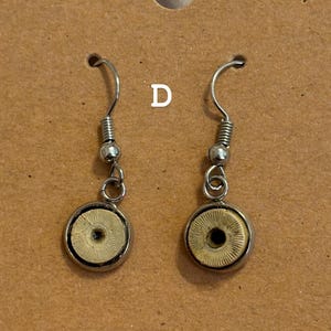 Puede incluir: Un par de pendientes colgantes con ganchos plateados y peque&ntilde;os colgantes circulares en tonos beige y plateado. Los pendientes se muestran en una tarjeta marr&oacute;n con la letra "D" impresa.