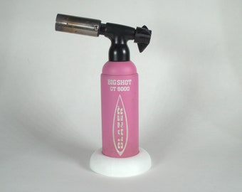 May include: A pink torch lighter with a black handle and a silver nozzle. The lighter is labeled "BIG SHOT GT 8000" and has a white base.
